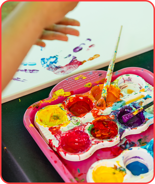 Child's hand holds a colorful paintbrush and palette, celebrating creativity at a vibrant pride event.